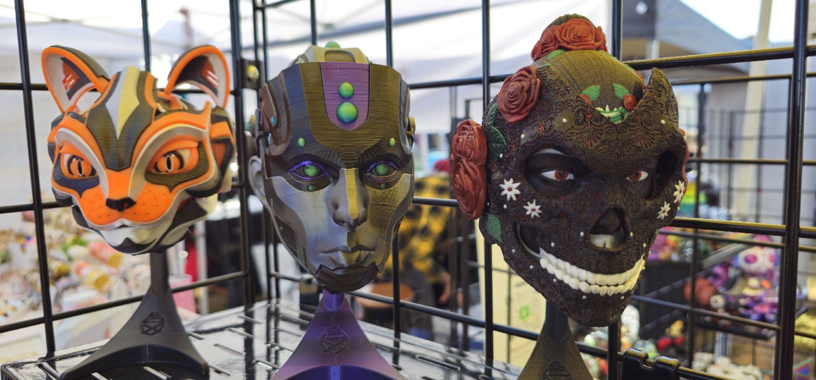 Orange cat mask, cyborg face and rose-covered sugar skull on display stands