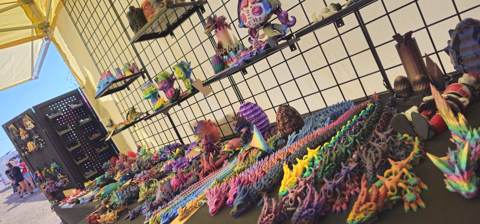 Outdoor tent booth with a long table of rainbow articulated dragons, eggs and shelves of colorful prints