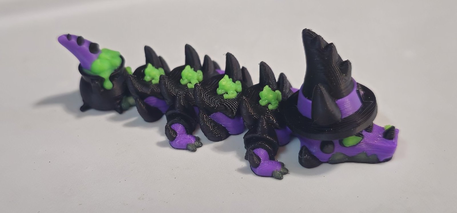 Purple and black articulated witch dragon with a pointed witch hat, a bubbling cauldron tail and tiny green frogs along its back